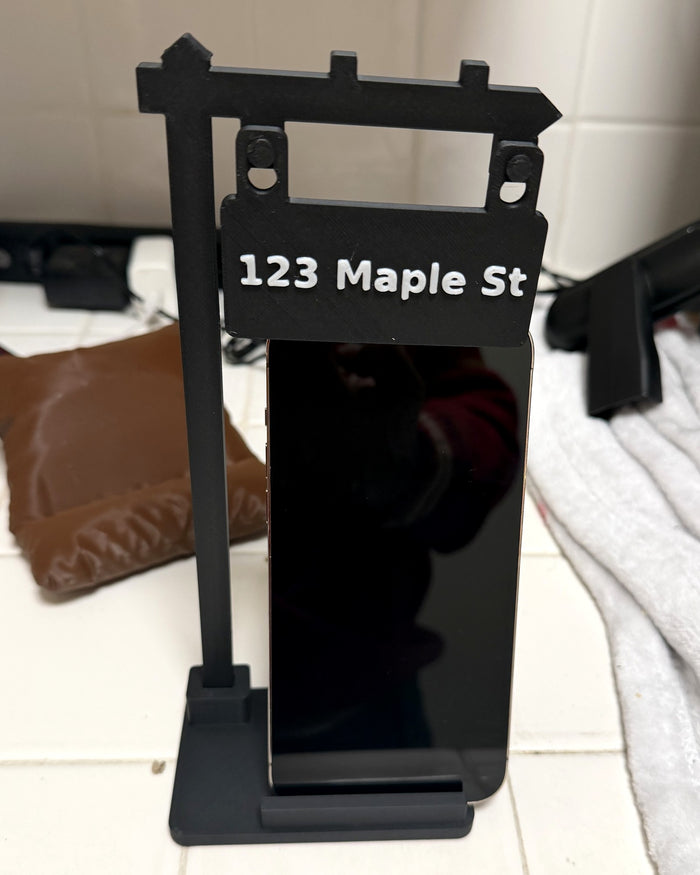 Real photo: black SOLD sign phone stand with white 123 Maple St address sign, holding a phone