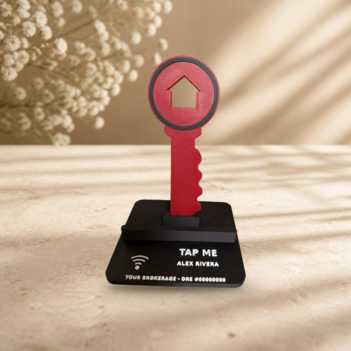 Key Phone Stand with a red house key on a black base with white TAP ME text, on a cream stone surface (real product photo on a styled background)