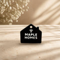 Black NFC house keychain with white raised MAPLE HOMES lettering and tap icon on a cream stone surface (real product photo on a styled background; key ring not included)