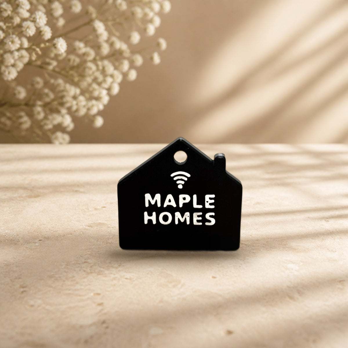 Black NFC house keychain with white raised MAPLE HOMES lettering and tap icon on a cream stone surface (real product photo on a styled background; key ring not included)