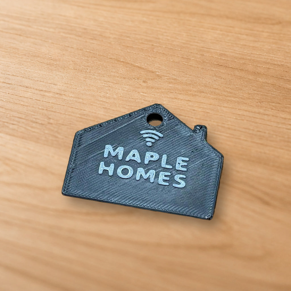 Close-up flat lay of the real NFC house keychain showing raised MAPLE HOMES lettering, tap icon, ring hole and printed layer texture on a wood surface